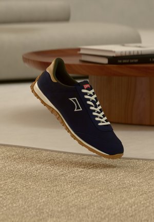 Baskets bleu marine en daim avec lacets blancs et logo blanc. Elles ont une semelle en caoutchouc avec un motif alvéolé et des accents en cuir beige sur le talon.