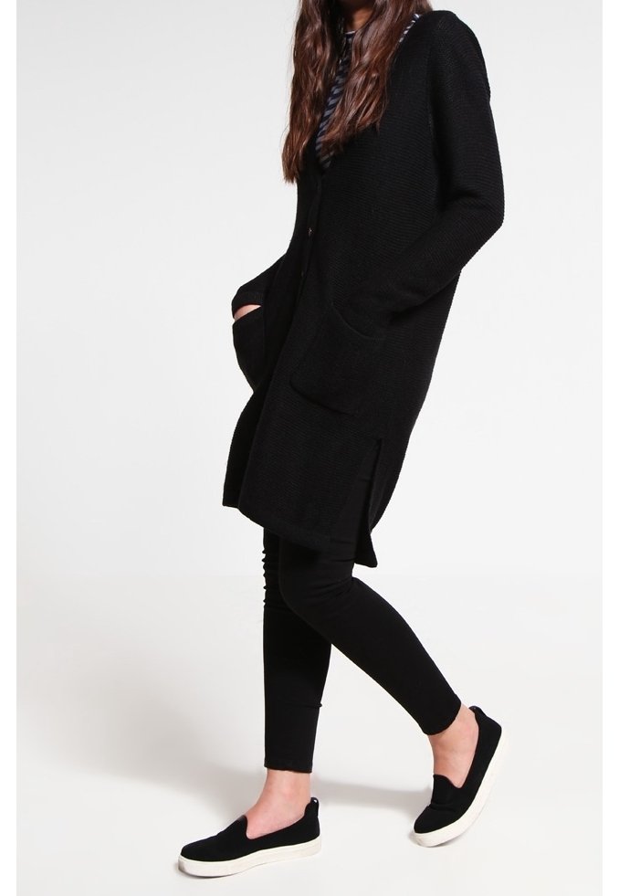 Femme portant un long cardigan noir, des leggings noirs et des chaussures sans lacets noires avec des semelles blanches, debout devant un fond clair uni.