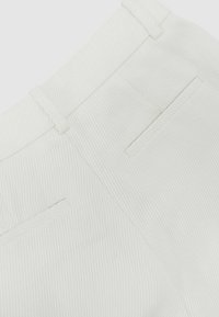 Primer plano de pantalones de tela blanca texturizada que muestran la pretina, las trabillas y dos bolsillos traseros de ojal.
