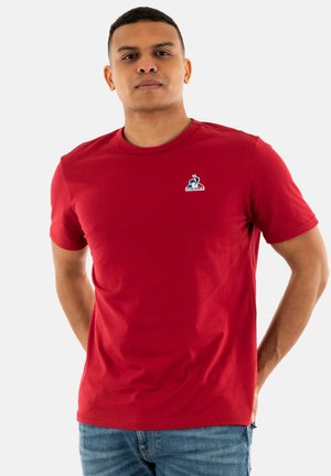 Red cotton t-shirt with short sleeves, round neckline, and small blue logo on the left chest. Smooth texture, casual fit.