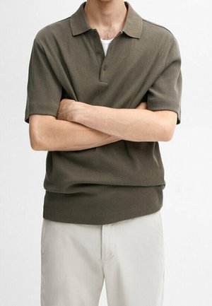 Man with arms crossed wearing olive green short-sleeve knit polo and light beige pants against plain white background.