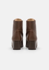 Un par de botas de cuero marrón hasta el tobillo con tacones gruesos, mostradas desde la parte posterior sobre un fondo blanco.