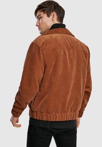 Giacca in velluto a coste marrone, caratterizzata da texture a coste, colletto e polsini elasticizzati. Il design posteriore include una cucitura a yoke e una fascia in vita sagomata.