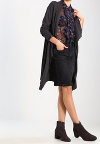 Gonna di denim nera, sovrapposta a una blusa floreale trasparente blu navy e a un lungo cardigan grigio. Abbinata a stivali scuri alte fino alla caviglia.