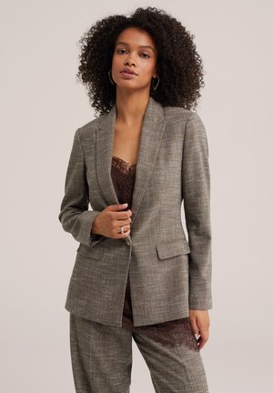 Donna con capelli ricci che indossa un blazer grigio a quadri e pantaloni abbinati sopra una maglia di pizzo marrone scuro, in piedi davanti a uno sfondo neutro.