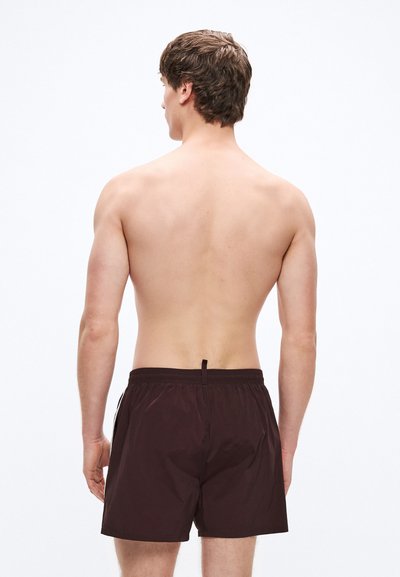 Pantalones cortos de baño marrones con una superficie texturizada, que cuentan con una cintura elástica y un pequeño lazo en la parte trasera. Vista desde atrás.