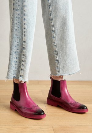 Magentowe skórzane botki Chelsea z czarnym czubkiem, elastycznymi bocznymi panelami i dekoracyjnymi perforacjami, w połączeniu z postrzępionymi jasnymi dżinsami.