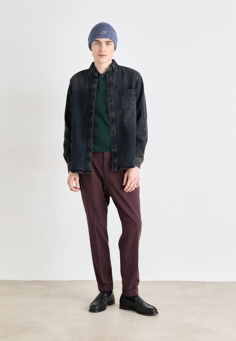 Chemise en denim foncé, polo vert, pantalons bordeaux, bonnet en tricot gris, chaussures noires. Design simple avec des boutons et une poche avant sur la chemise.