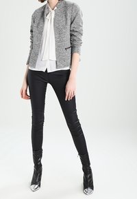 Giubbotto bomber grigio texturizzato, blouse bianca con fiocco, pantaloni neri aderenti e stivaletti neri alla caviglia con punte metalliche.