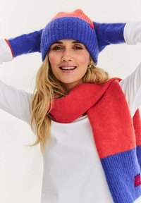 Dziany czapka typu beanie w kolorach fioletowym i koralowym, charakteryzująca się grubą prążkowaną fakturą. Pasujący szalik w kolorach koralowym i fioletowym owinięty wokół szyi.