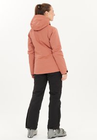 Vesimproof vaaleanpunainen takki hupulla, yhdistettynä mustiin housuihin ja harmaisiin hiihtokengiin. Oma erityinen muotoilu ja sileä rakenne.