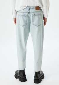 Jeans de mezclilla de color azul claro con un ajuste relajado, que cuentan con dos bolsillos traseros y una etiqueta de cuero con la marca en la cintura. Llevados con botas negras.