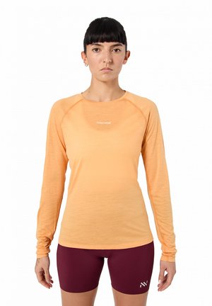 Donna che indossa una maglia arancione a maniche lunghe e pantaloncini da ciclismo marroni in piedi contro uno sfondo bianco.