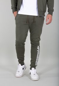 Pantaloni jogger verde oliva realizzati in morbido materiale con design affusolato, caratterizzati da scritte bianche sulla gamba sinistra e vita regolabile con cordino.