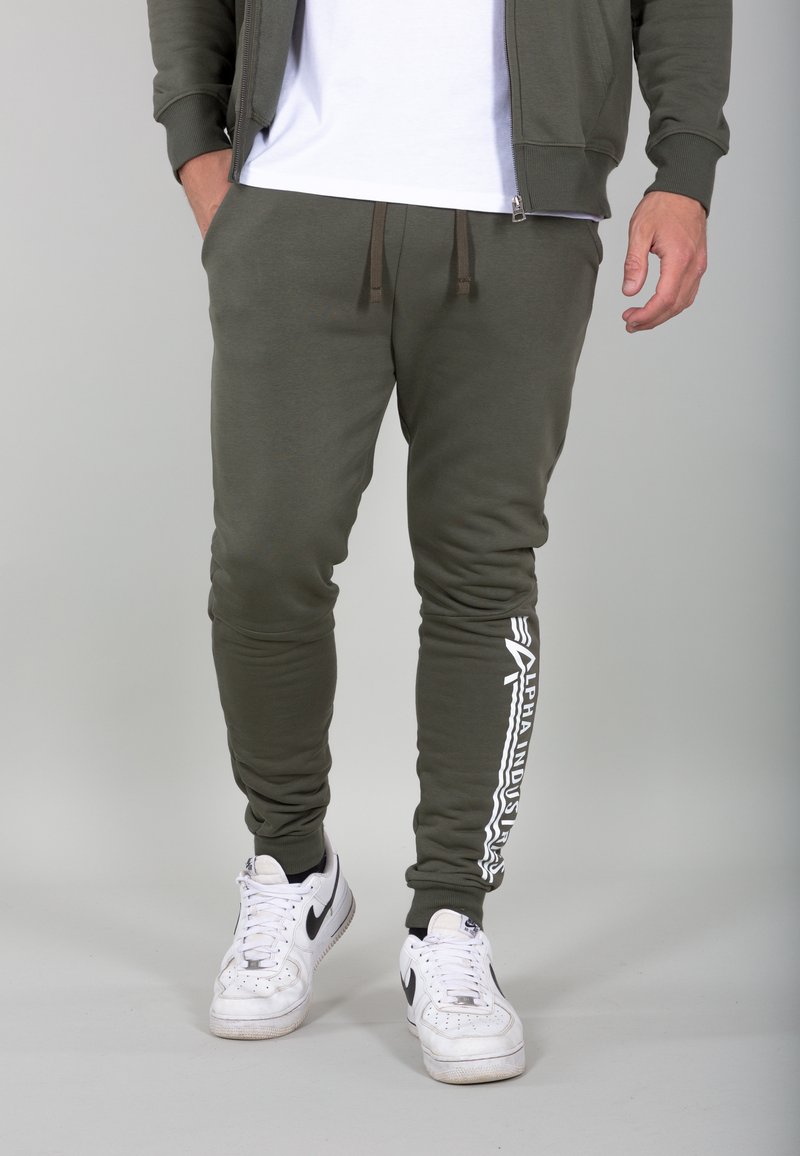 Pantaloni jogger verde oliva realizzati in morbido materiale con design affusolato, caratterizzati da scritte bianche sulla gamba sinistra e vita regolabile con cordino.