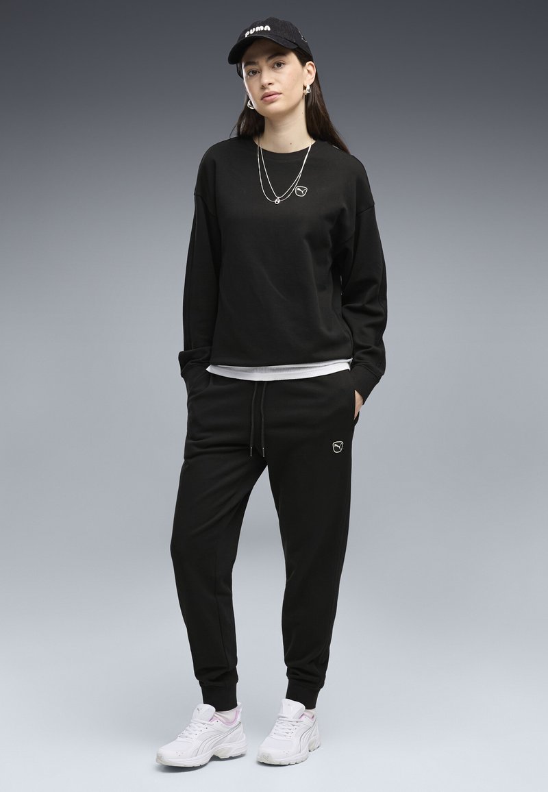 Jeune femme en sweat-shirt noir, jogging et casquette Puma, portant des baskets blanches et des colliers argentés, debout les mains dans les poches.