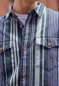 Camisa de botones con rayas verticales en verde, marino y blanco. Hecha de una tela texturizada con dos bolsillos en el pecho y cuello redondeado.