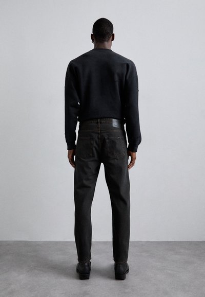 Musta sviiter pikkae varrukatega, tumedad teksad ja mustad kingad, vaadeldud seljalt ühetaolise tausta ees.