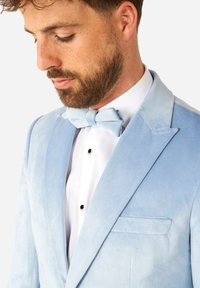 Chaqueta de esmoquin de terciopelo azul claro con solapas de muesca, un bolsillo y un pajarita a juego sobre una camisa blanca con botones negros.