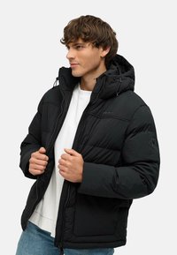 Giacca nera imbottita con chiusura a zip e cappuccio. Presenta una texture trapuntata e tasche laterali. Indossata sopra una camicia bianca e jeans di denim.