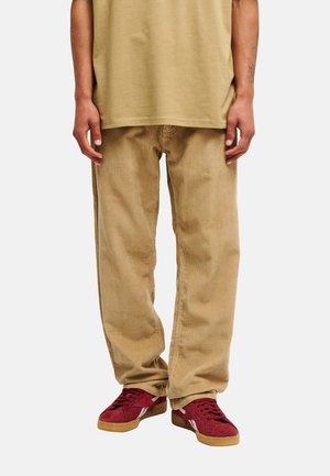 Personne portant un pantalon en velours côtelé beige, une chemise kaki et des baskets rouges à semelles en gomme, debout devant un fond uni.