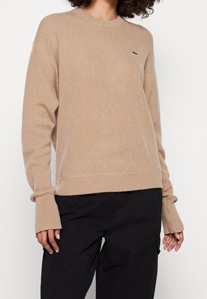 Osoba ubrana w beżowy sweter z długim rękawem z małym logo na piersi oraz czarne spodnie, stojąca na jednolitym białym tle.