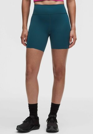 Person, der bærer turkis højttaljede sportsshorts, lyserød cropped top, sorte sokker og sorte løbesko, står på en ensfarvet baggrund.