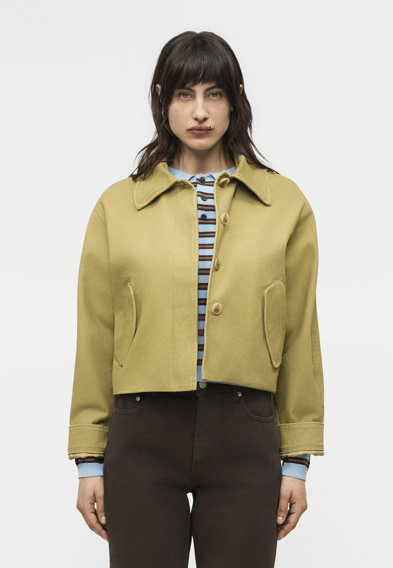 Γυναίκα που φοράει κοντό μπουφάν σε μουσταρδί χρώμα με κουμπιά πάνω από μπλε και καφέ ριγέ πουκάμισο και σκούρο παντελόνι, στέκεται μπροστά σε απλό φόντο.