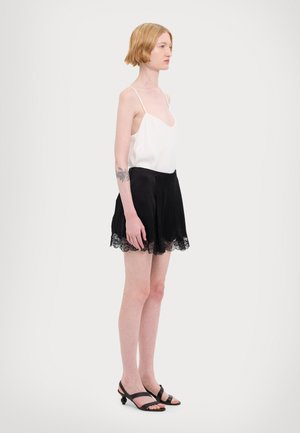 Witte satijnen camisole met dunne bandjes, gecombineerd met zwarte satijnen shorts met een kanten zoom. Model draagt zwarte sandalen met bandjes.