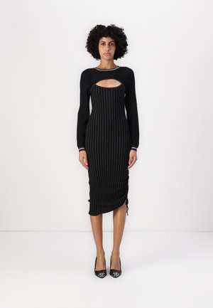 Robe noire à côtes avec des manches longues, comportant une découpe à l'avant, des accents argentés et un détail froncé au niveau de l'ourlet. Portée avec des talons noirs.