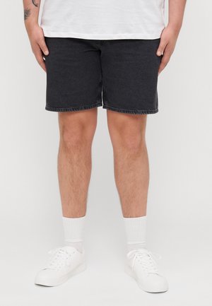 Jambes d'une personne portant un short en jean noir, des chaussettes blanches et des baskets blanches, debout devant un fond clair uni.