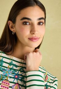 Bílo-zelený pruhovaný svetr s barevně vyšitými vánočními ozdobami. Modelka má dlouhé tmavé vlasy a nosí malé kruhové náušnice.