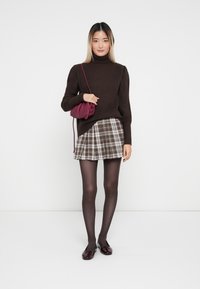 Smeđi rebrasti pulover s turtleneck ovratnikom, karirana A-linija suknja, prozirne crne čarape, ljubičasta torba i sjajne crne balerinke. Svijetla pozadina.