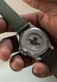 Rustfrit ståluhr med en grøn stofrem. Bagsiden har teksten "EXPEDITION NORTH TIMEX" samt et minimalistisk design med teksturerede detaljer.