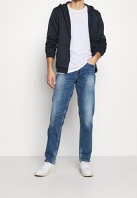 Marineblå hættetrøje med lynlås, hvid T-shirt, lyseblå jeans og hvide sneakers. Hættetrøjen har snore ved hætten og forlommer.