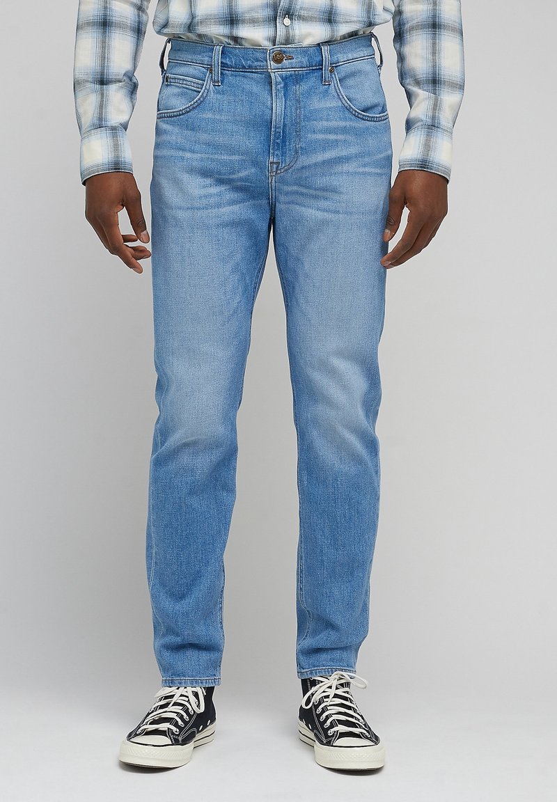 Personne portant un jean bleu clair, une chemise à carreaux boutonnée et des baskets en toile noires, debout face à un fond uni.