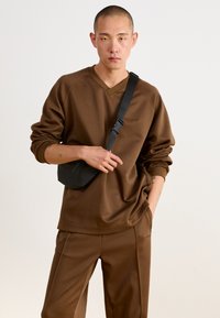 Sweatshirt marron avec un col en V, une coupe décontractée et des manches longues, associé à un pantalon assorti. Tient un petit sac bandoulière noir.