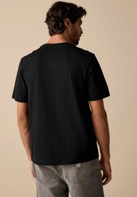 Camiseta de algodón negra de manga corta, con cuello redondo y textura suave, mostrada desde la parte posterior sobre un fondo beige.
