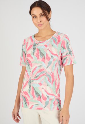 Vrouw draagt een shirt met korte mouwen in pastelroze, groen en blauw met een abstract bladmotief en crème broek tegen een effen lichte achtergrond.