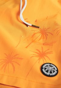 Shorts de bain orange avec un motif de palmiers, dotés d'un cordon blanc et d'un patch rond noir. Gouttes d'eau visibles sur le tissu.