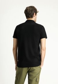 Camisa polo negra de manga corta, material de algodón, corte recto, sin logotipos ni patrones visibles, llevada con pantalones cargo de color oliva.