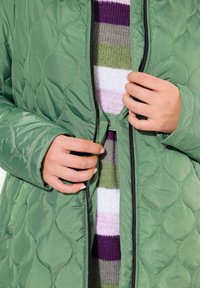 Prošívaná zelená bunda se zapínáním na zip; má lesklou texturu, kulaté vzory a černé kovové prvky; vrstvená přes pruhovaný svetr.