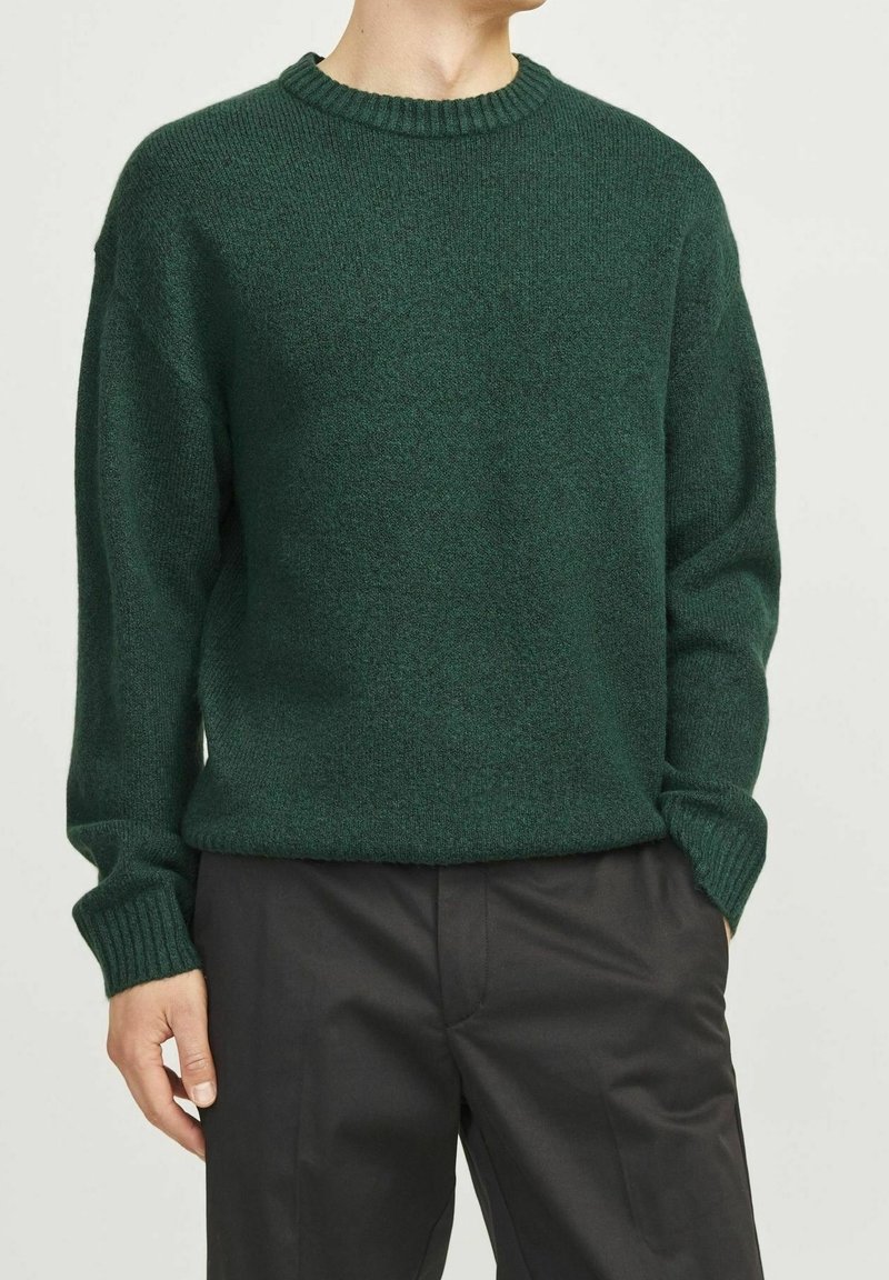 Dunkelgrüner, grob gestrickter Pullover mit einem gerippten Rundhalsausschnitt und langen Ärmeln, kombiniert mit schwarzen Hosen. Weicher Stoff und lässige Passform.