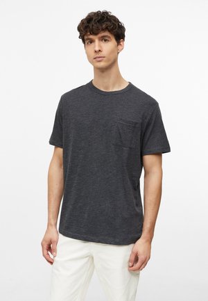Jeune homme aux cheveux bouclés portant un T-shirt gris foncé à manches courtes avec une poche poitrine et un pantalon blanc, debout devant un fond uni.