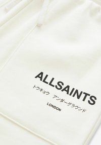 Hvid sweatshirt med en frontlomme, snørehætte og sort tekst, der læser "ALLSAINTS," "トウキョウ," og "LONDON." Glat tekstur.