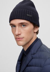 Beanie a coste di colore blu navy con un piccolo tag con logo. Il modello indossa un giubbotto imbottito scuro sotto. Messa a fuoco sul design e sulla texture del cappello.