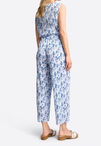 Mouwloze jumpsuit van lichtblauwe stof met een bloemenpatroon. Beschikt over een elastische taille en wijde broekspijpen. Gecombineerd met witte sandalen.