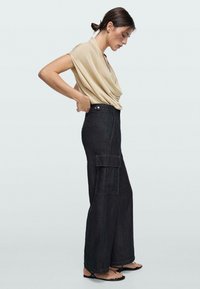 Femme ajustant un pantalon cargo noir taille haute, portant un haut beige drapé à manches courtes et des sandales noires à lanières, debout de profil.