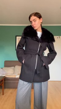 Melns mētelis ar mākslīgās kažokādas apkakli, rāvējslēdzēja aizdari un jostu viduklī, ar sānu kabatām un piegriezumu. Zem tā pelēkas platām kājām bikses.