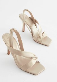 Beige High Heelsandalen mit viereckigen Zehen, weichen Lederriemen, die vorne überkreuzt sind, und einem gerafften Fersenriemen für zusätzliche Details.